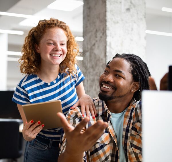A white woman looking at a laptop having a conversation with a black man sitting on a chair in front of her
