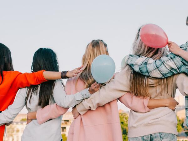Four women in bright clothing holding each other's waist and shoulder and looking up to the sky