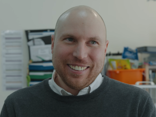 A smiling person with a shaved head and trimmed beard, wearing a white collared shirt under a dark sweater, looking slightly to the side while indoors in an office setting.