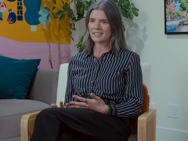 A woman with long, grayish-brown hair sits in a chair indoors, wearing a black and white striped blouse and black pants, speaking and gesturing with her hands. Colorful artwork and a plant are visible in the background.