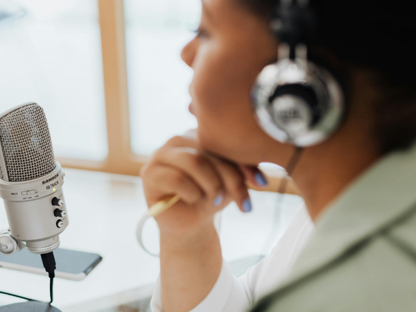 A woman wearing over-ear headphones sits at a desk, positioned beside a microphone on a stand. They are recording a podcast.