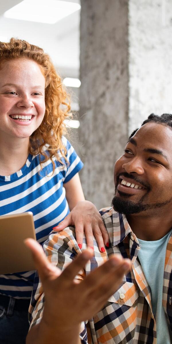 A white woman looking at a laptop having a conversation with a black man sitting on a chair in front of her