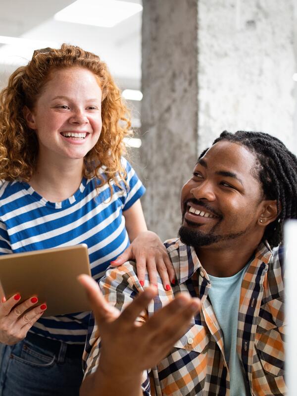 A white woman looking at a laptop having a conversation with a black man sitting on a chair in front of her