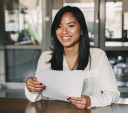 Young smiling Asian woman in a white blouse sitting at the desk holding pieces of paper