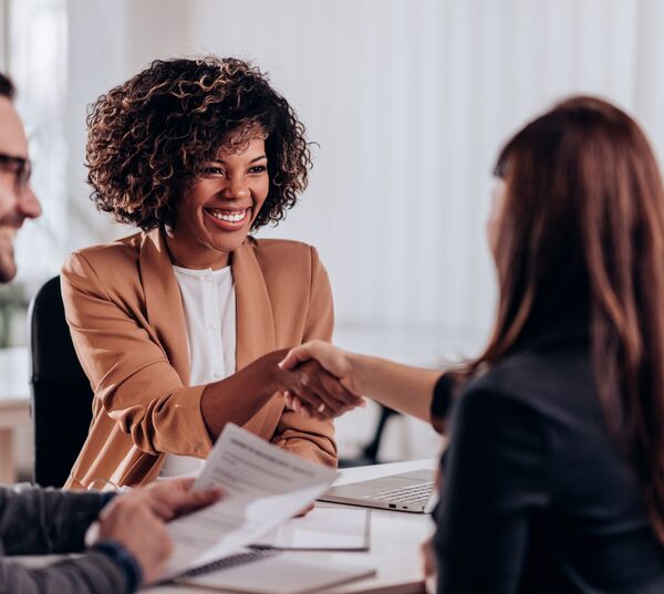 A joyful black woman sitting at the desk shaking hands with another woman 