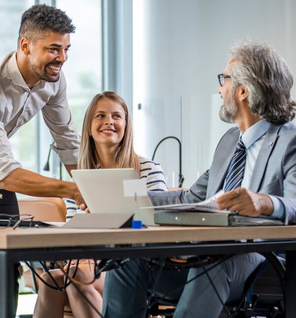 A black man and white woman  smiling at a white man sitting at the table with a laptop and binder 