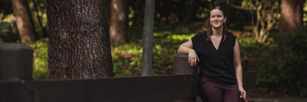 White woman leaning against the fence with a blurred background of a large tree trunk