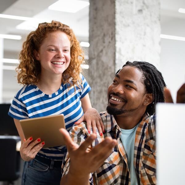 A white woman looking at a laptop having a conversation with a black man sitting on a chair in front of her